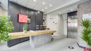 Un espace d’accueil avec un comptoir en bois, un menu numérique affiché sur un écran, une caméra de surveillance, un ascenseur et des plantes en pot.