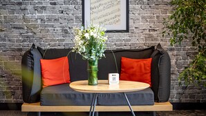 Une pièce moderne avec un mur en briques, un canapé avec des coussins orange, une table ronde en bois portant un code QR, et une partition musicale encadrée accrochée au mur.