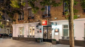 L’entrée d’un hôtel, avec une enseigne indiquant « ibis Paris Daumessin Porte Dorée », se distingue par sa façade traditionnelle en briques, ses nombreuses fenêtres et ses espaces de détente en plein air.