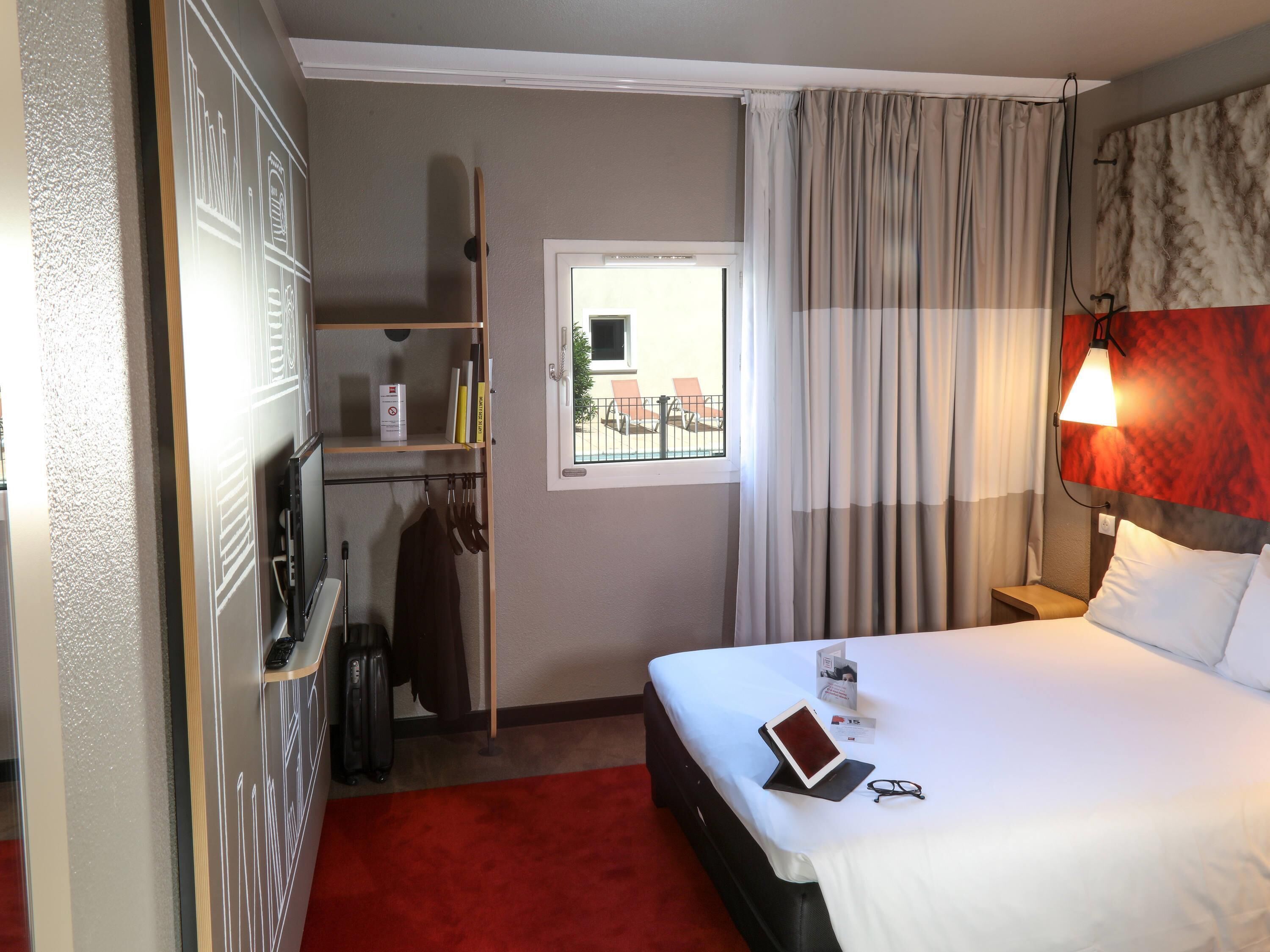 A hotel room with a bed, a bedside table, a lamp, a window with curtains, a wardrobe, and a red carpet.