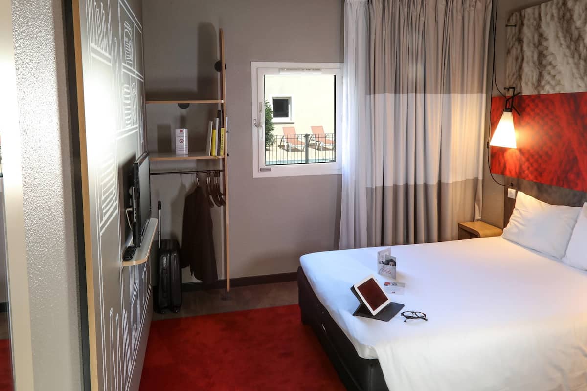 A hotel room with a bed, a bedside table, a lamp, a window with curtains, a wardrobe, and a red carpet.