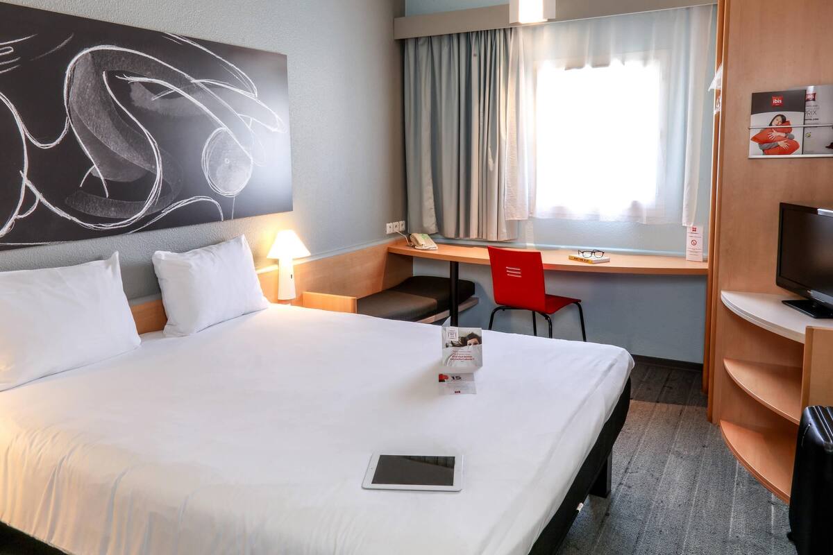 Habitación de hotel con cama, escritorio, silla, televisión y ventana con cortinas.