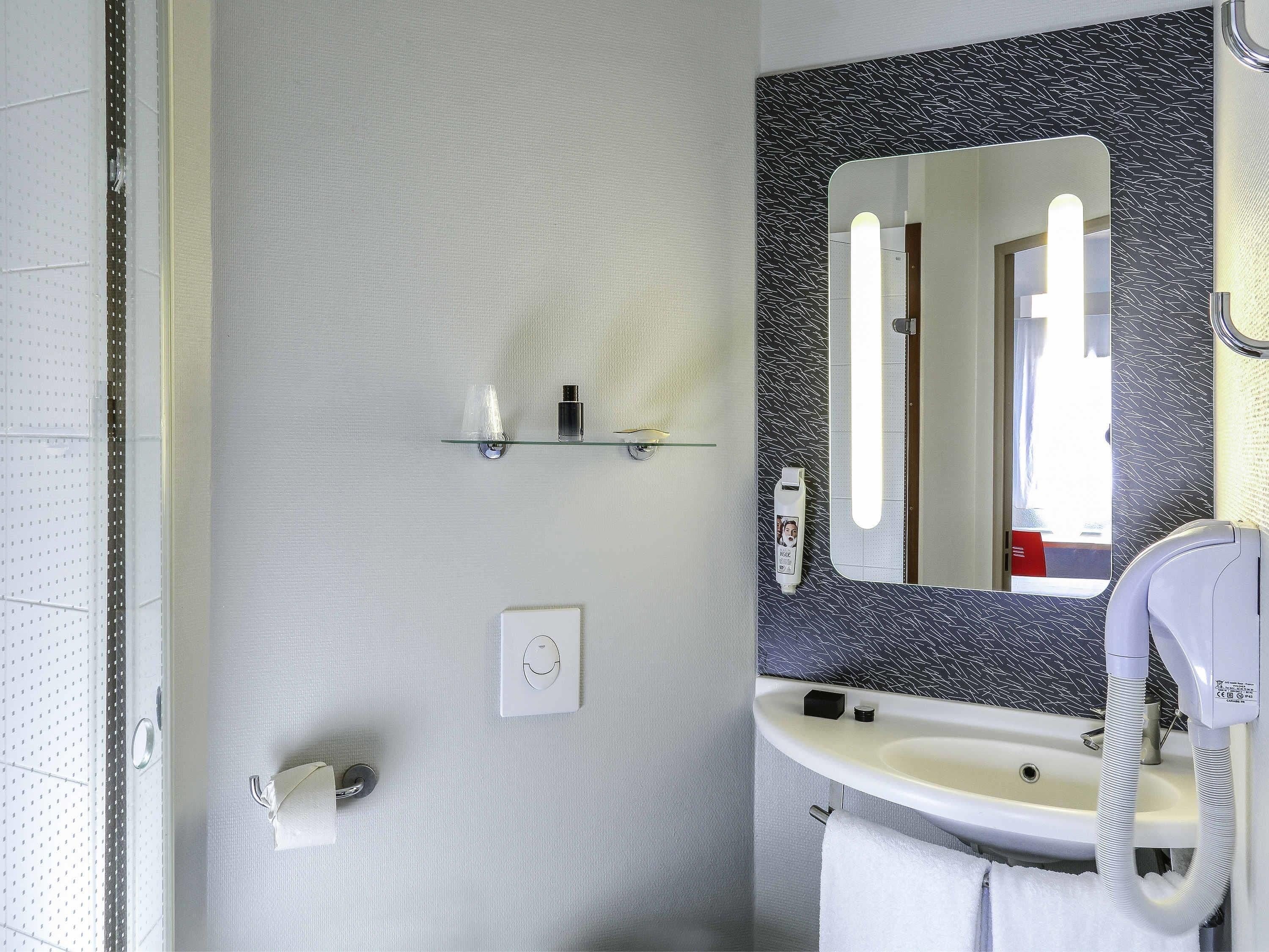 Un baño con un lavamanos blanco, un espejo con un respaldas negro estampado, un secador de pelo y una toalla colgada en la pared.