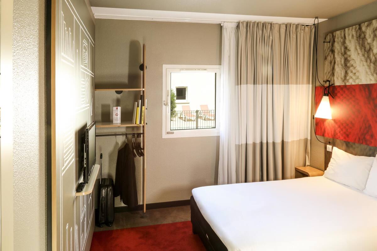Habitación de hotel con cama, escritorio, silla, armario y ventana con cortinas.