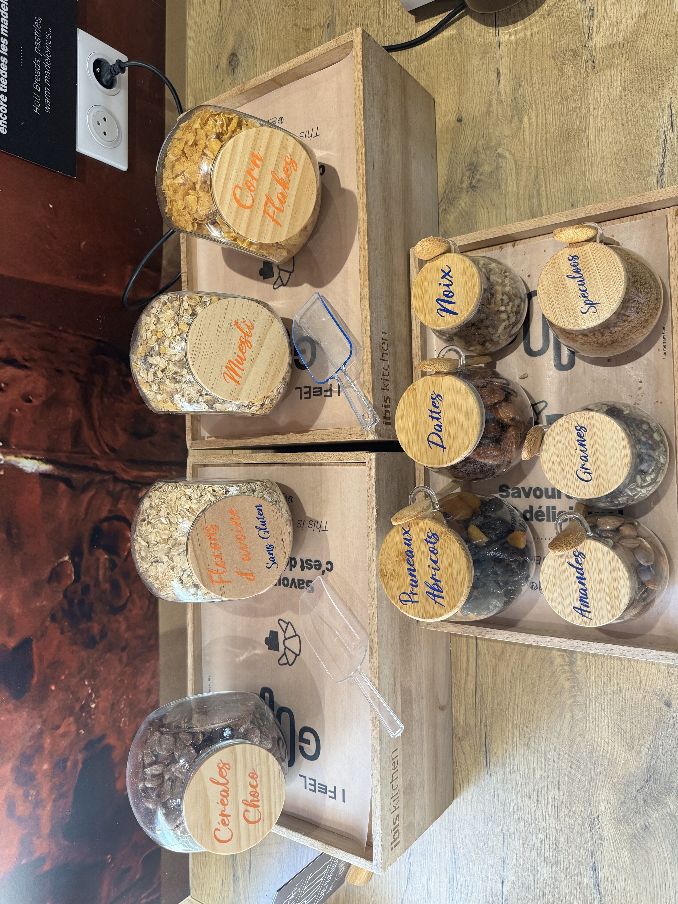 A wooden display with various food items labeled with names like "Corn Flakes," "Muesli," and "Pruneaux Abricots."