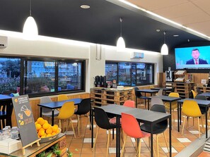 Interior de restaurante com mesas, cadeiras, uma tela de televisão e vista para o exterior.