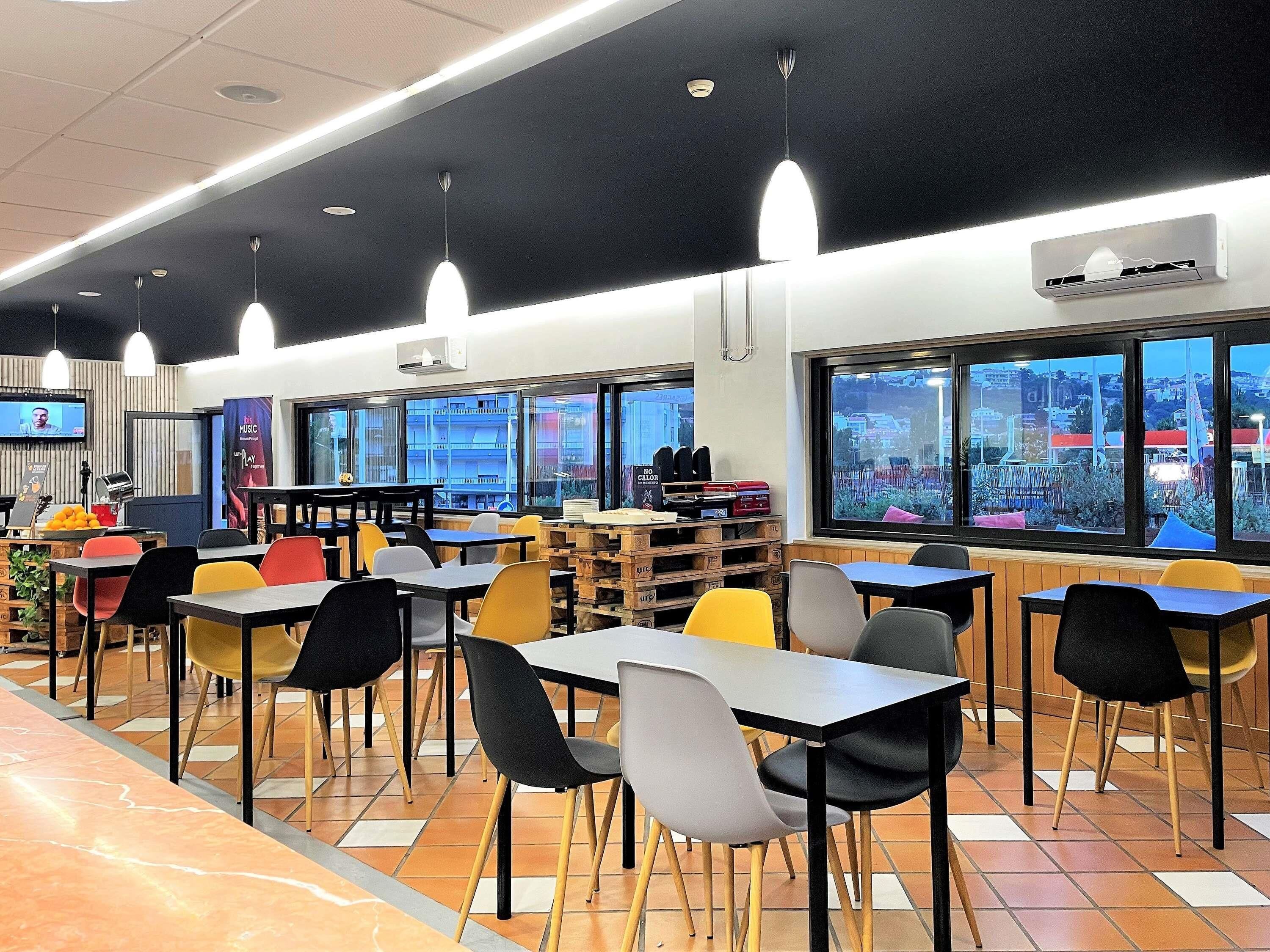 A modern cafeteria with multiple tables and chairs, large windows offering views of the city, and ceiling lights.