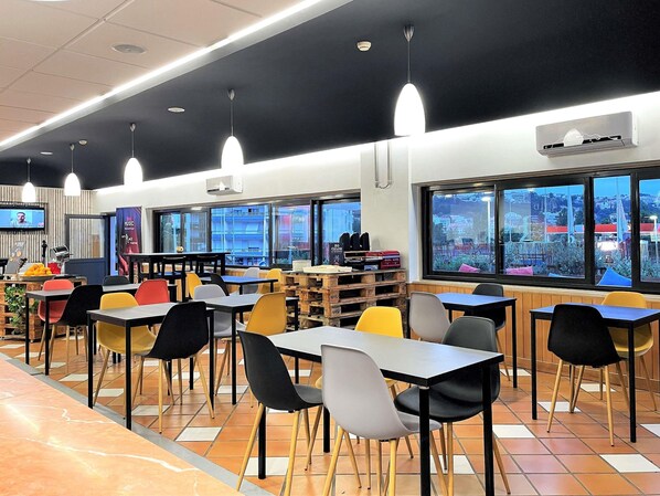 A modern cafeteria with multiple tables and chairs, large windows offering views of the city, and ceiling lights.
