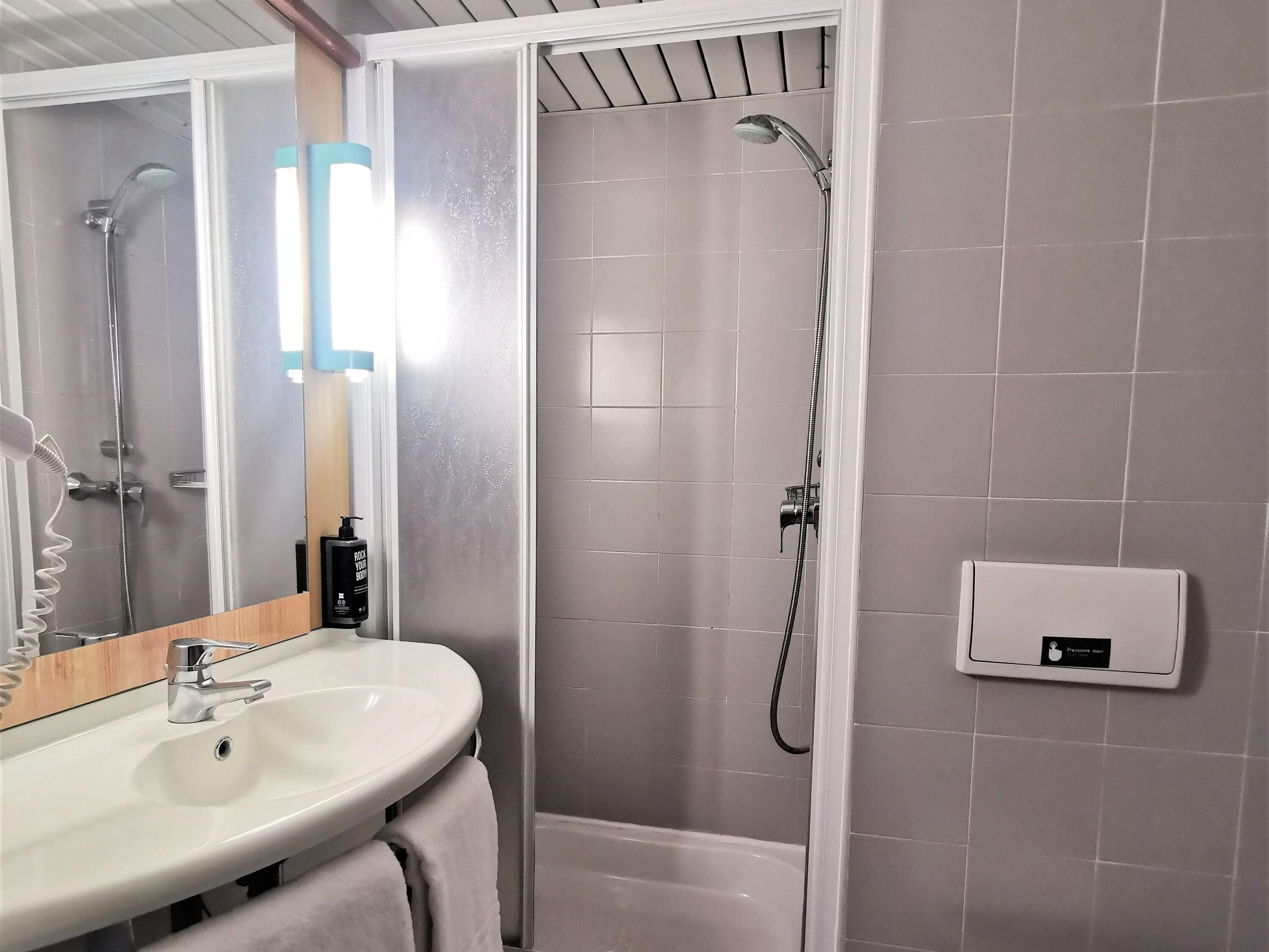 A bathroom with a white sink, a mirror, a shower with a handheld showerhead, and a control panel on the wall.