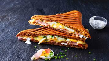 A grilled sandwich with visible layers of ham, cheese, and egg, accompanied by a small bowl of sauce and chopped herbs.
