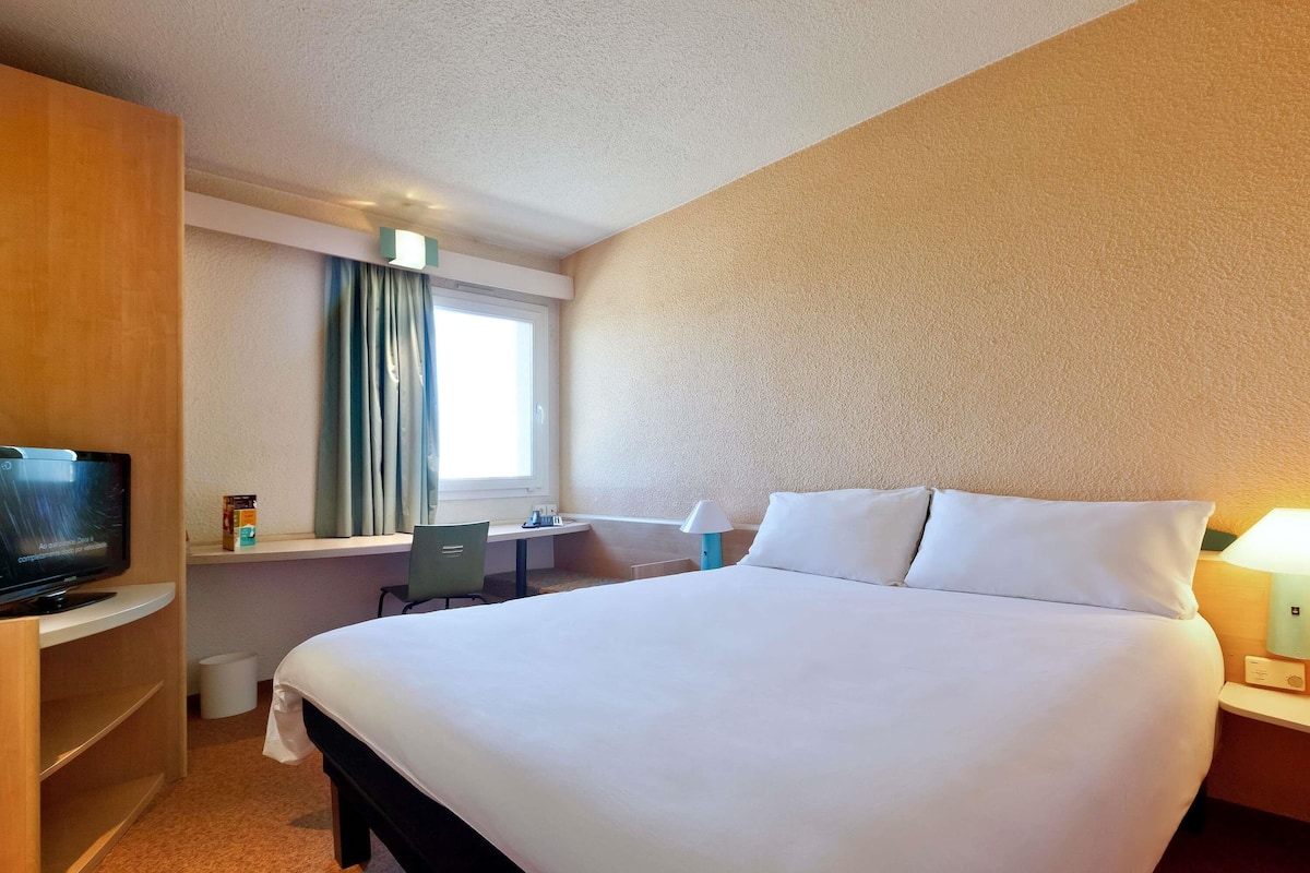A hotel room with a large bed, a desk with a chair, a television, and a window with curtains.