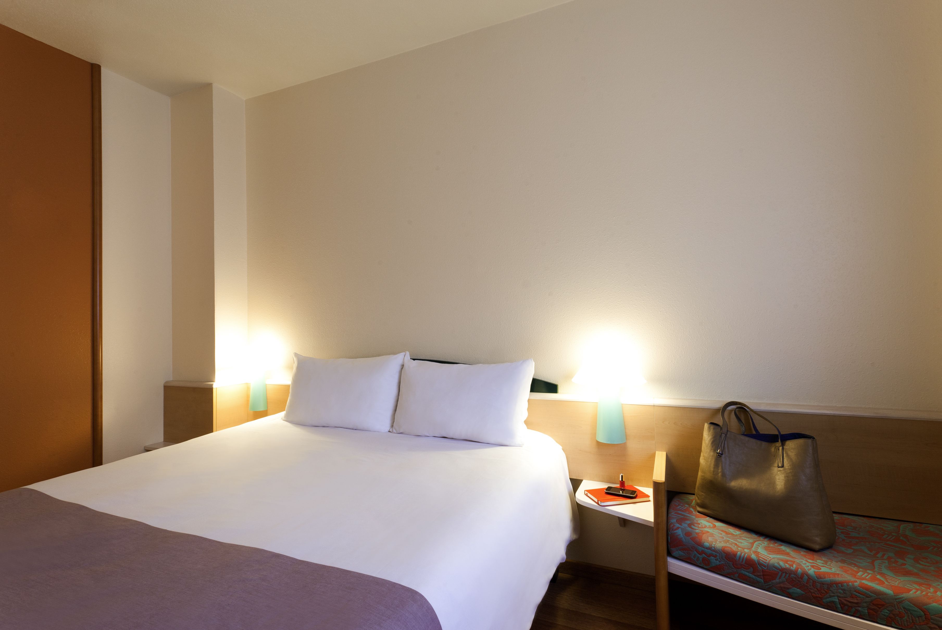 Una habitación de hotel con una cama grande, lámparas de noche, una mesita de noche y una silla con un bolso.