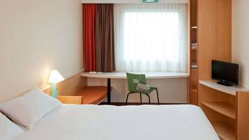 A hotel room with a bed, a desk with a chair, a television, and a window with curtains.