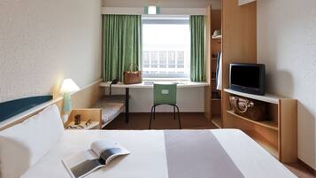 A hotel room with a bed, a desk with a chair, a television, and a window with curtains.