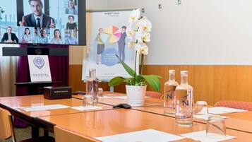 A meeting room with a large screen displaying a video conference, a Novotel logo, a poster with a pie chart, and a potted orchid.