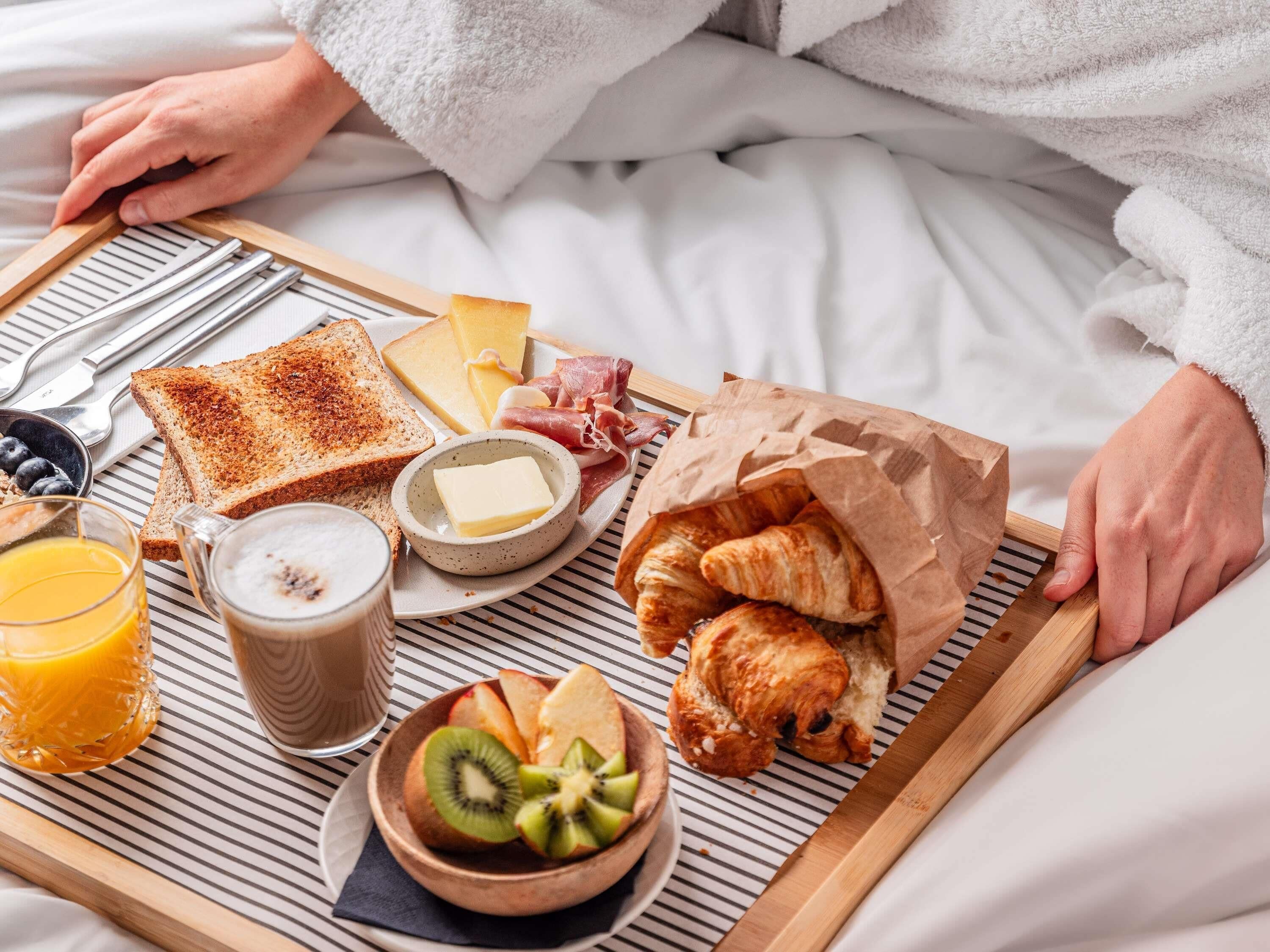 Bandeja de desayuno con tostadas, queso, jamón, cruasanes, fruta y jugo sobre una cama.