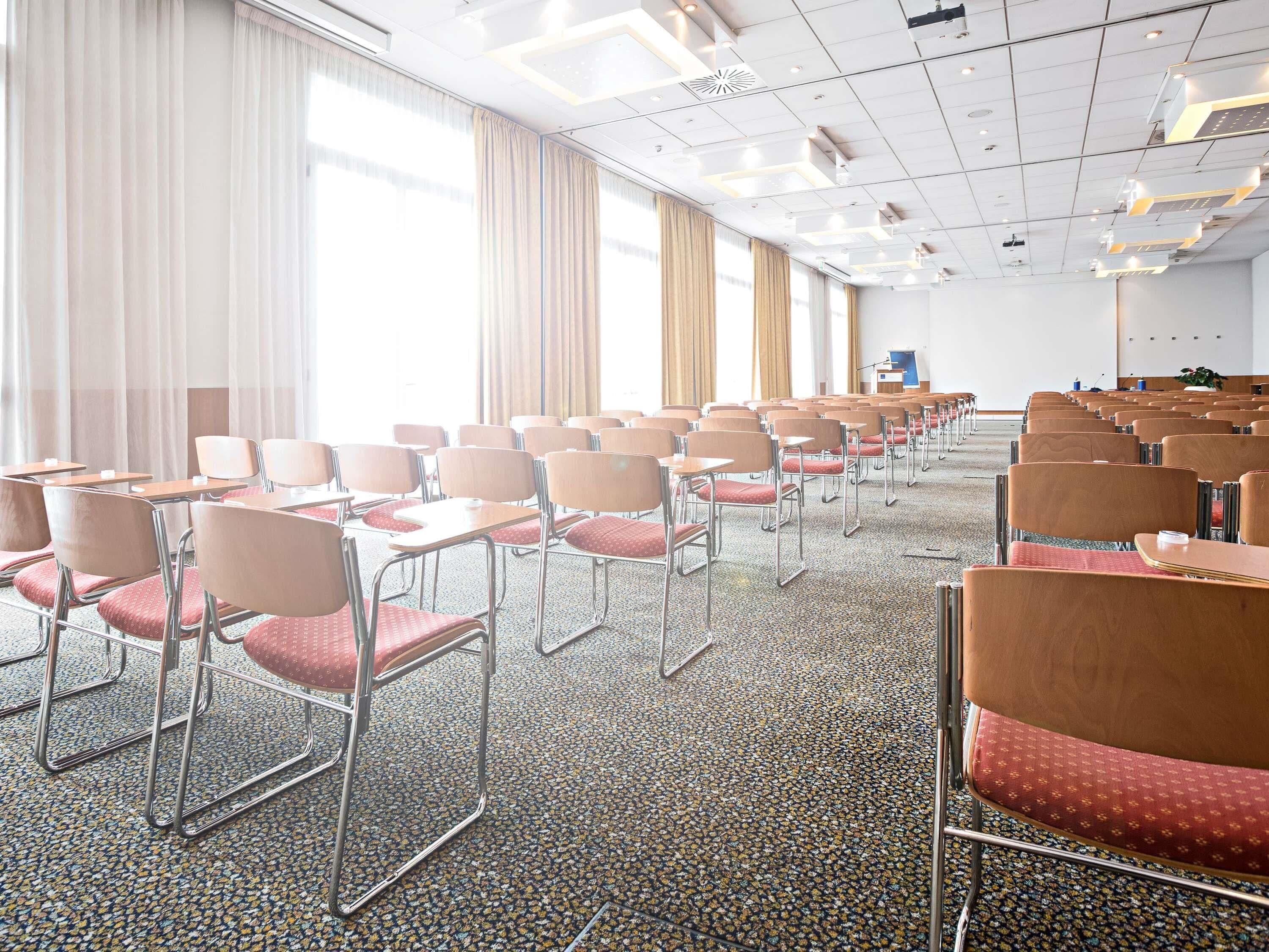 Un salón de conferencias con filas de sillas y mesas, ventanas amplias con cortinas y un techo con iluminación empotrada.