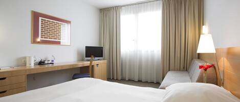 A hotel room with a large bed, a desk with a computer, a chair, and a window with curtains.