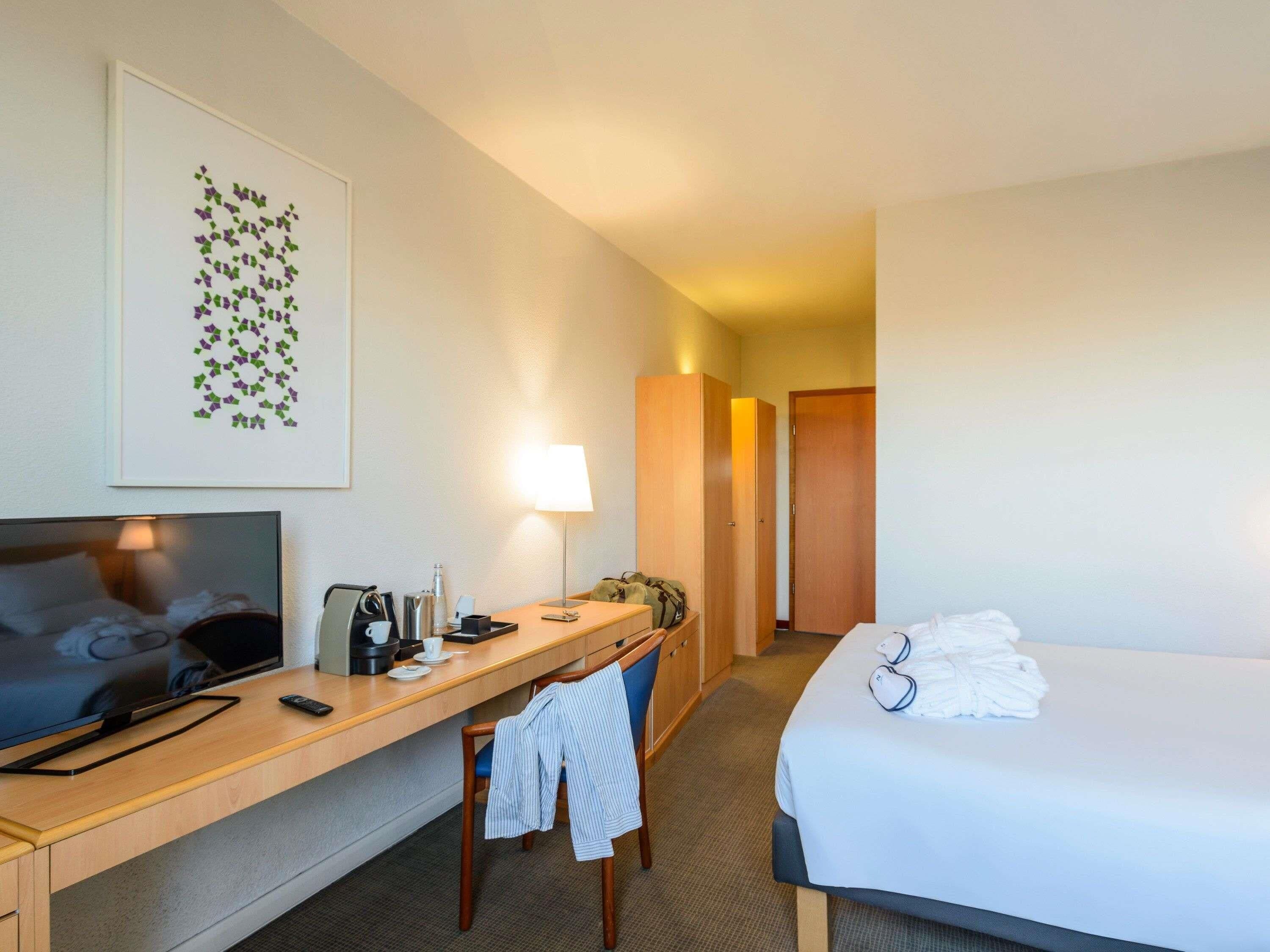 A hotel room with a bed, a desk with a chair, a TV, a lamp, and a framed picture on the wall.