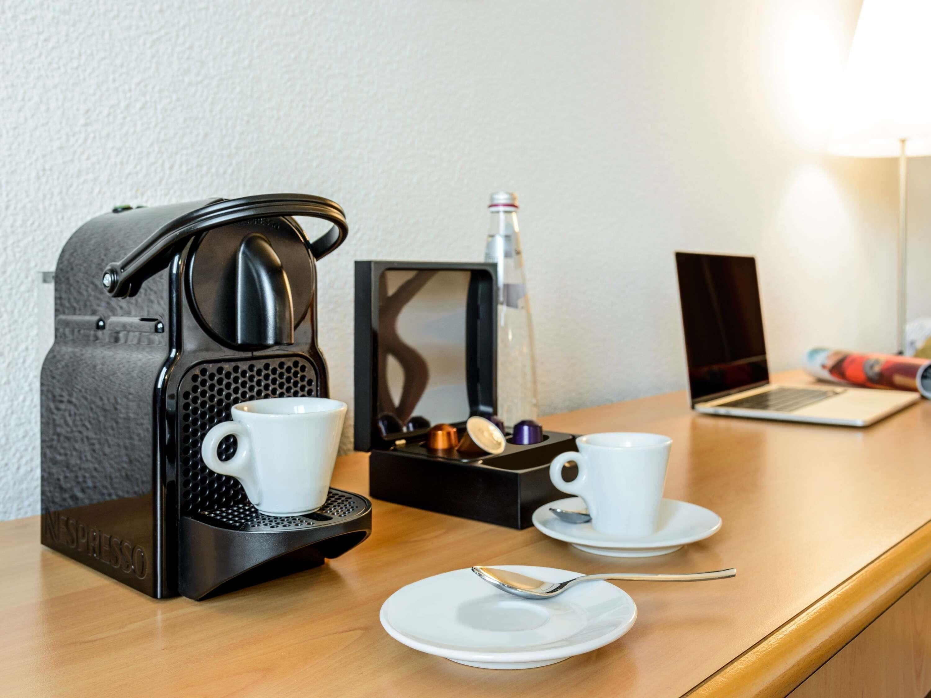 A coffee maker, two cups, a spoon, and a laptop on a wooden table.