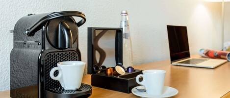 A coffee maker, two cups, a spoon, and a laptop on a wooden table.