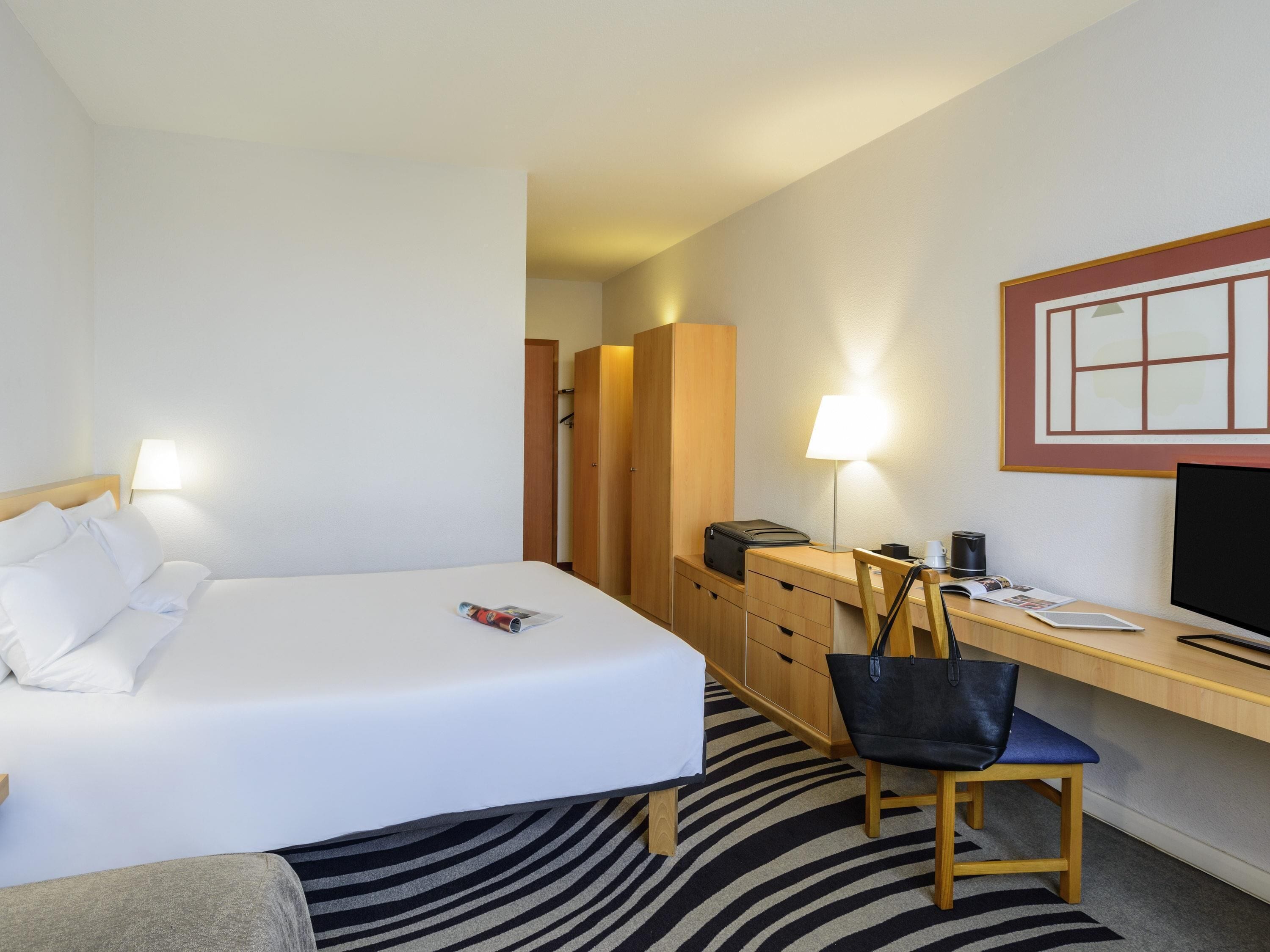 A hotel room with a bed, a desk, a chair, a lamp, a TV, and a wardrobe.