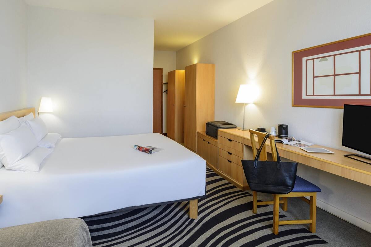 A hotel room with a bed, a desk, a chair, a lamp, a TV, and a wardrobe.