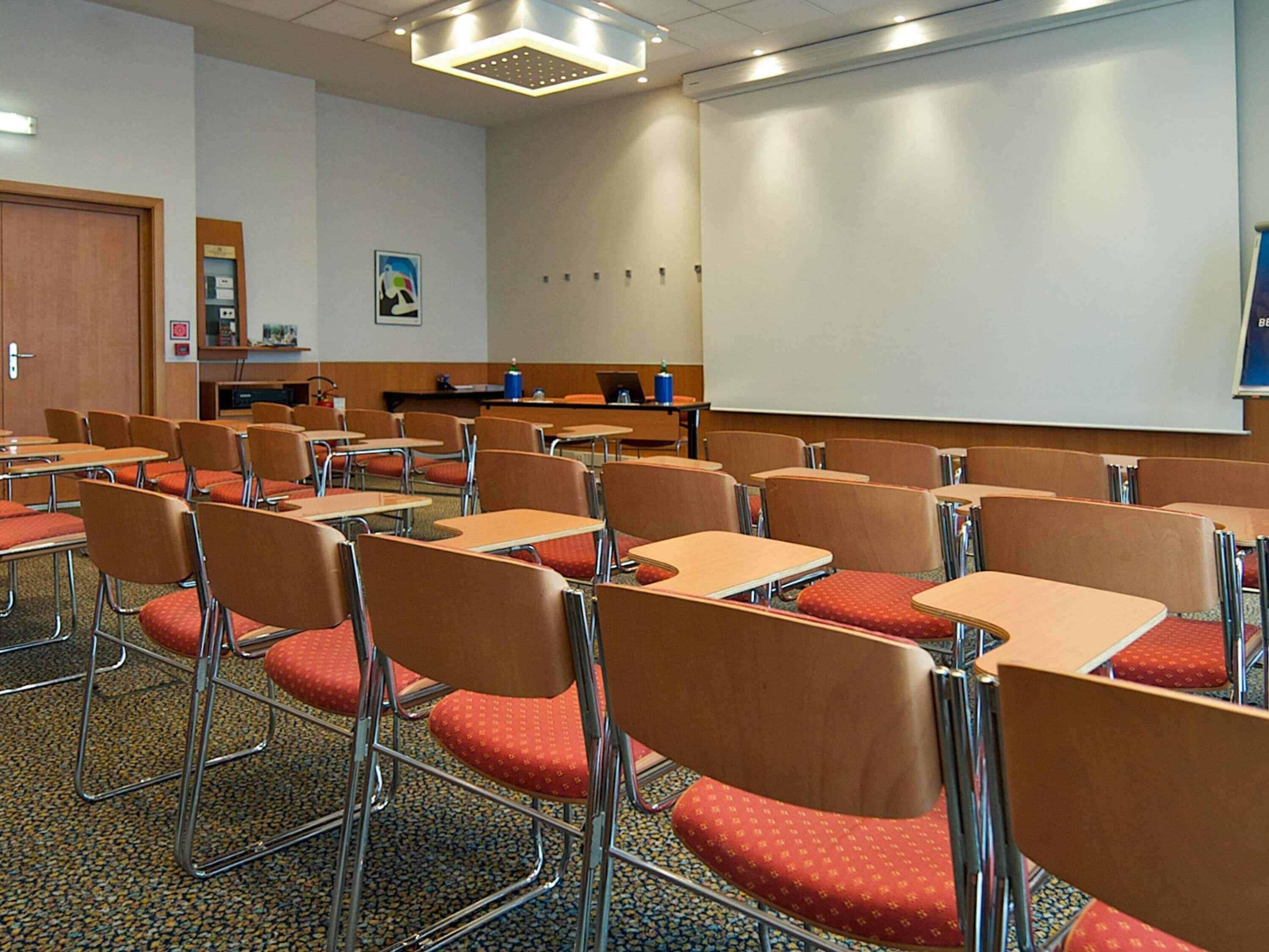 Un'aula con file di banchi e sedie, uno schermo per proiettore e una porta.