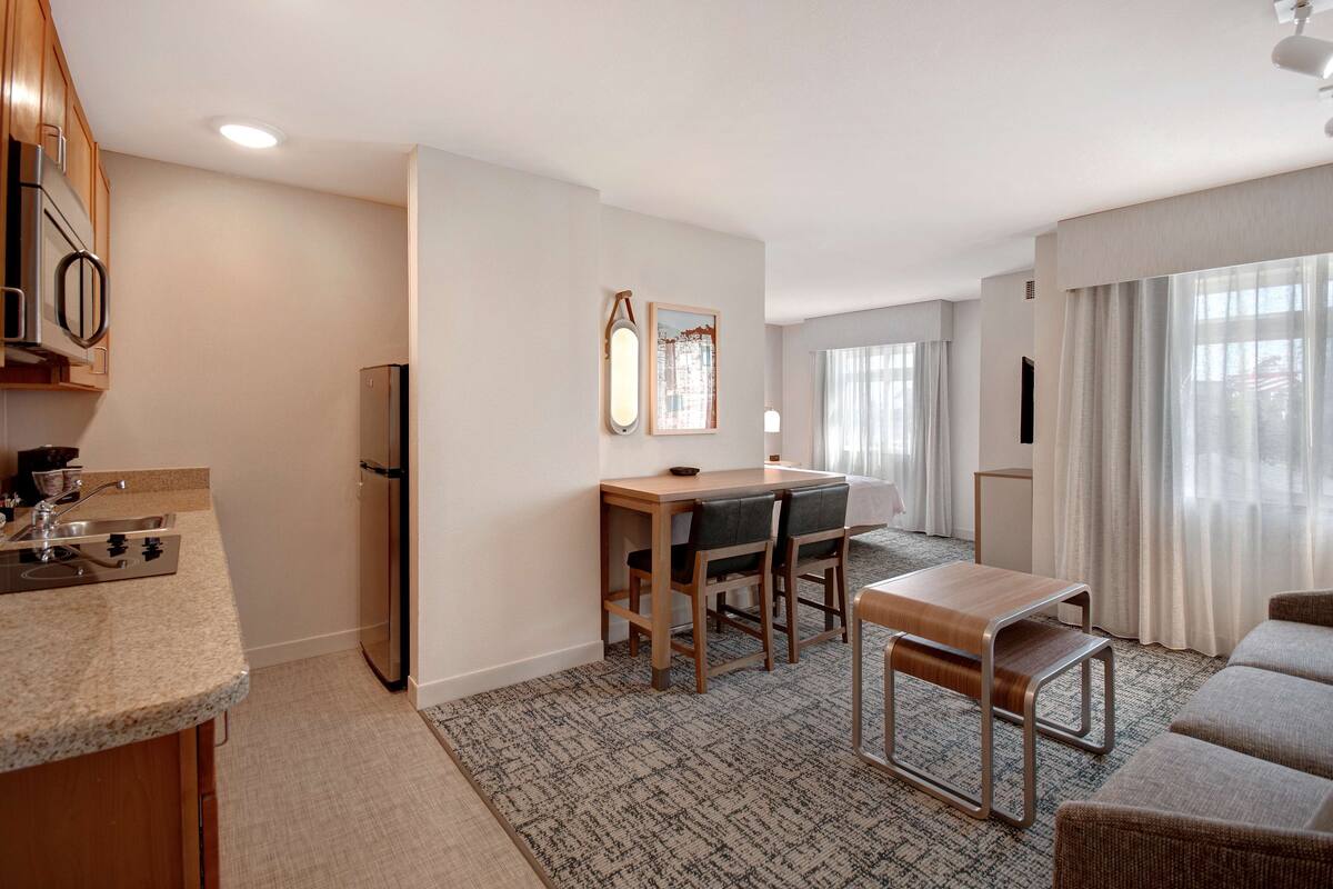 A modern kitchen with a built-in oven, a refrigerator, and a sink. There is a dining area with a table and chairs, and a sofa in the living area.