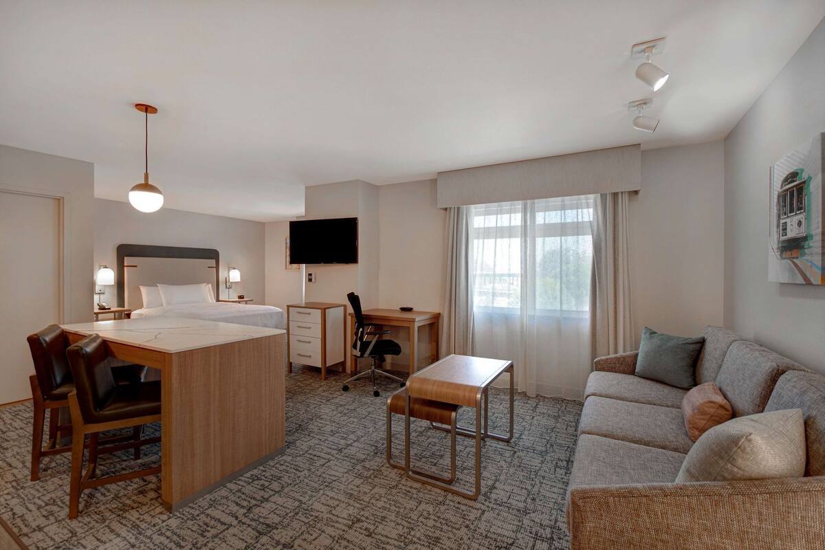 Habitación de hotel con cama, escritorio, silla, sofá, televisor y ventanas amplias.