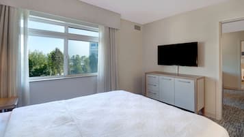 A hotel room with a large bed, a TV mounted on the wall, a window with curtains, and a view of trees and buildings outside.