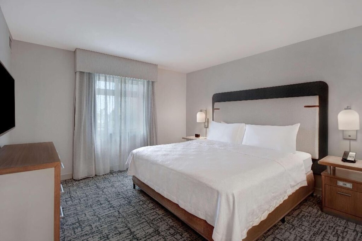 Habitación de hotel con una cama grande, mesitas de noche, televisor y ventana con cortinas.