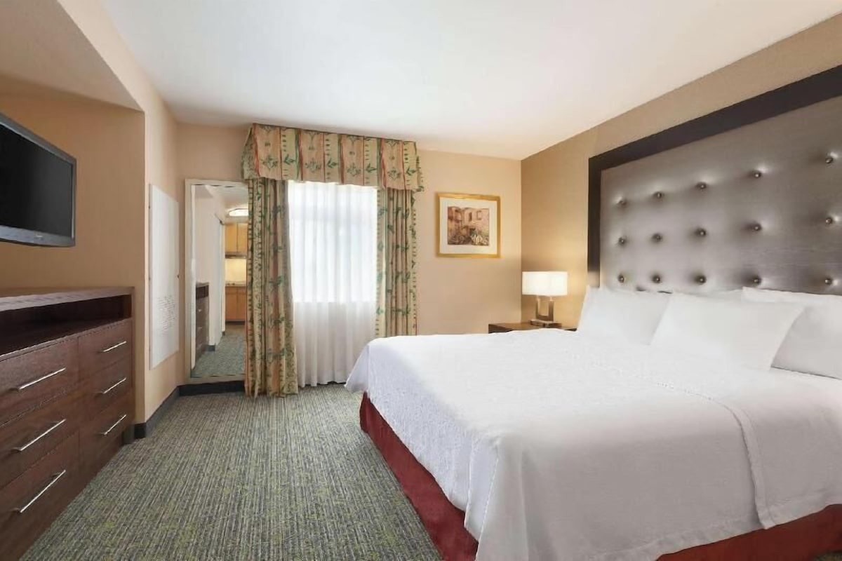Una habitación de hotel con una cama, televisión, cómoda, mesita de noche, lámpara y un cuadro en la pared.