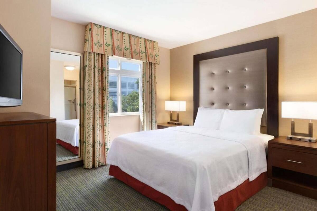 Habitación de hotel con una cama grande, dos mesitas de noche con lámparas, un televisor de pantalla plana, un espejo y una ventana con cortinas.