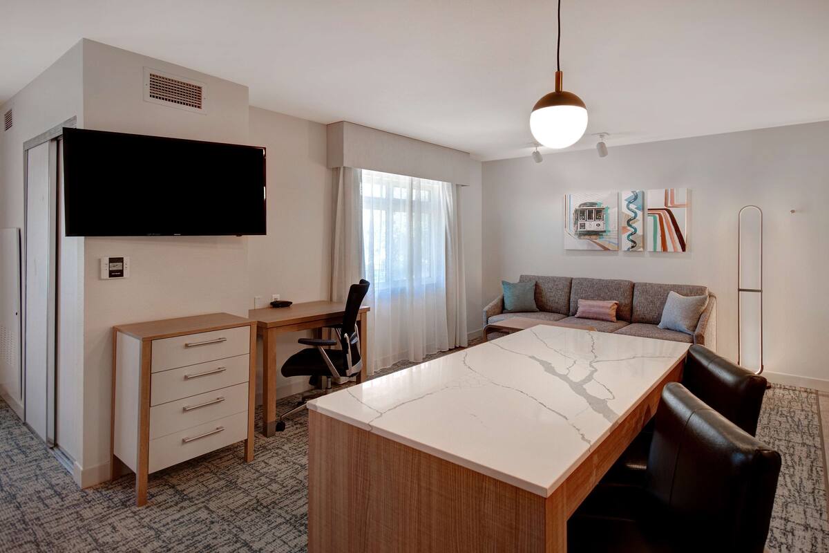 A modern hotel room with a large flat-screen TV, a wooden desk with drawers, a marble dining table, a sofa with cushions, and a wall-mounted artwork.