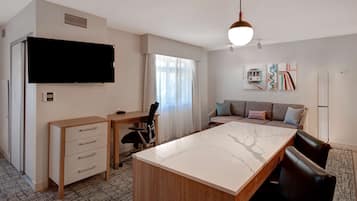 A modern hotel room with a large flat-screen TV, a wooden desk with drawers, a marble dining table, a sofa with cushions, and a wall-mounted artwork.