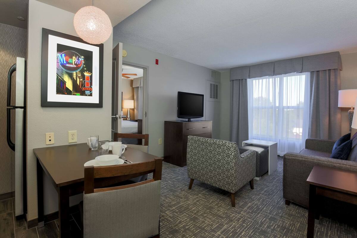 Habitación de hotel con comedor, sofá, sillón estampado, televisión y un amplio ventanal con cortinas.