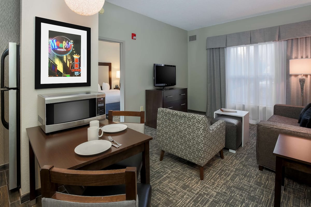 Habitación de hotel con zona de comedor, televisor, sillón estampado y un cuadro en la pared.