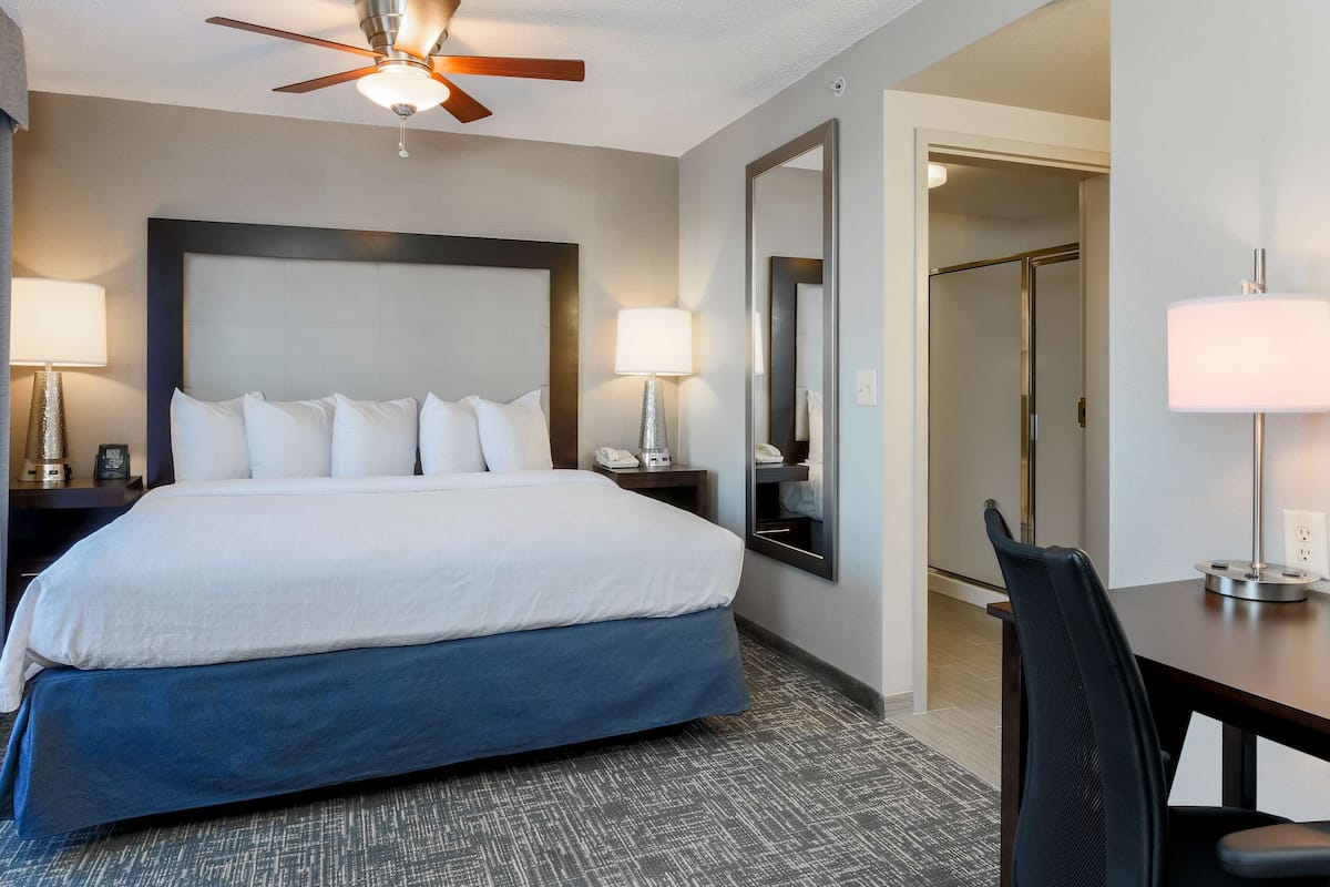 Una habitación de hotel con una cama grande, dos lámparas de noche, un ventilador de techo, un escritorio con una silla y un espejo.