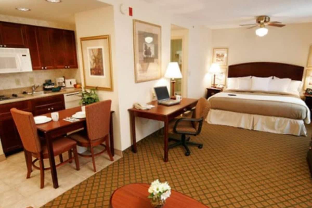 Habitación de hotel con cama, escritorio, silla, kitchenette y ventilador de techo.