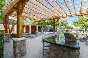 An outdoor patio area with a wooden pergola, stone pillars, a barbecue grill, and outdoor seating.