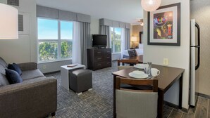 A hotel room with a sofa, a small table with a chair, a television, a framed picture on the wall, and a window with curtains. - Homewood Suites Southwind - Hacks Cross (Memphis)