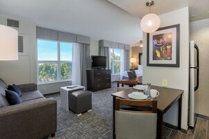 A hotel room with a sofa, a small table with a chair, a television, a framed picture on the wall, and a window with curtains.