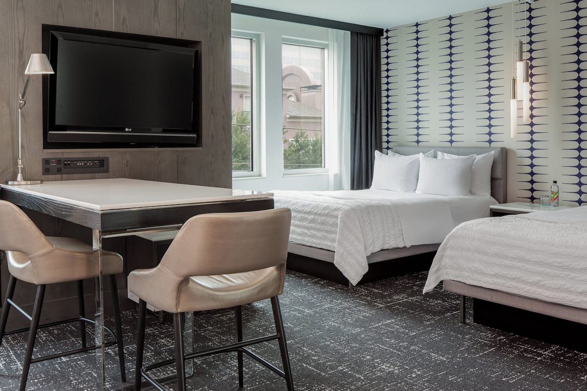 A hotel room with a bed, a desk with two chairs, a TV, and a window with curtains.