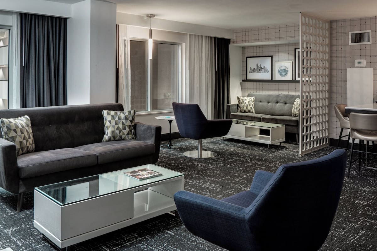 A modern hotel room with a grey sofa, a blue armchair, a glass coffee table, and a white side table. There is a dining area with a bar and a white cabinet.