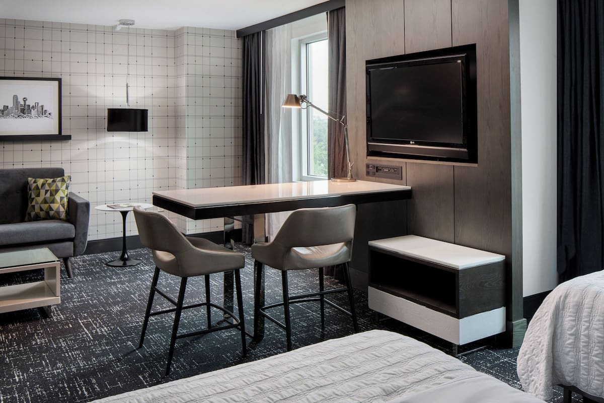 A hotel room with a bed, a desk with two chairs, a TV, a framed picture, and a window with curtains.