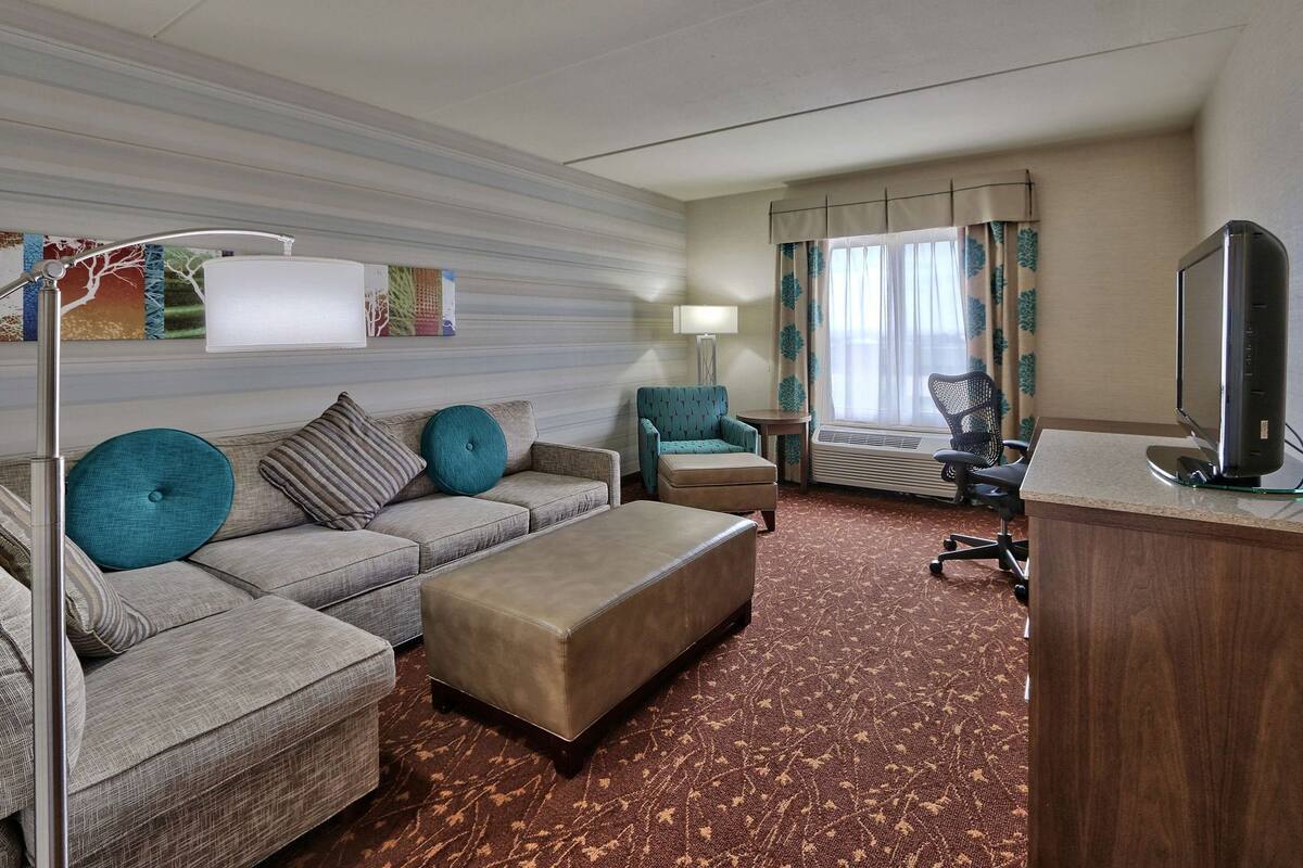 A hotel room with a sofa, ottoman, armchair, desk, and television.
