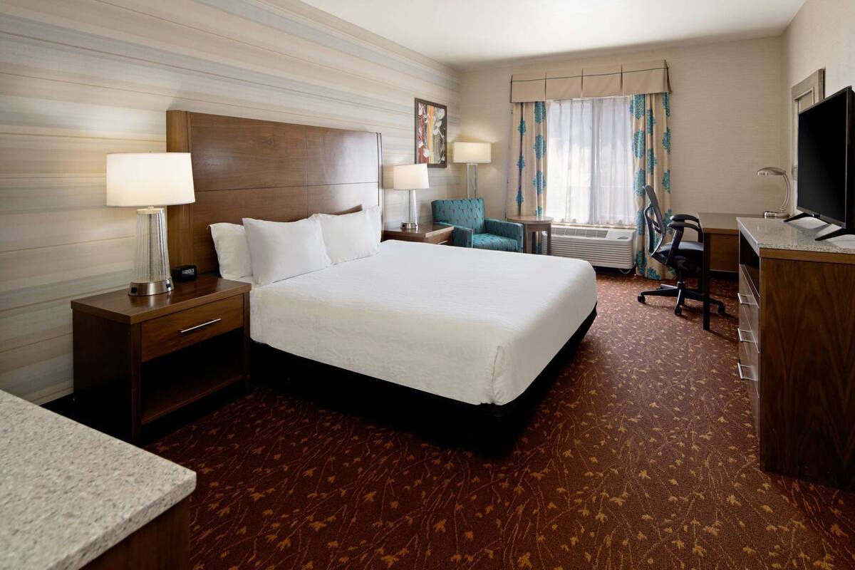 A hotel room with a large bed, a desk, a chair, a TV, and a lamp.