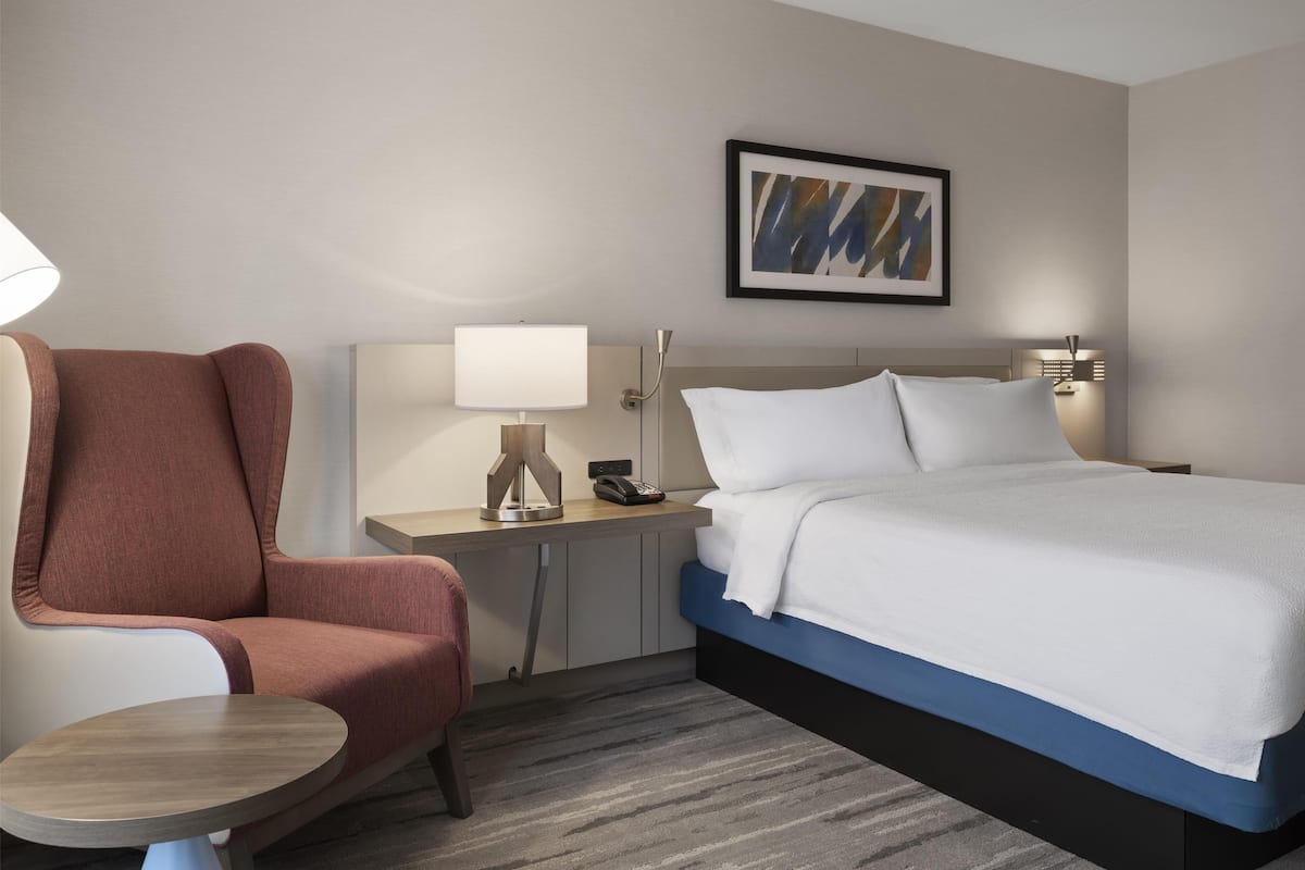 A hotel room with a bed, a desk with a lamp, a chair, and a painting on the wall.