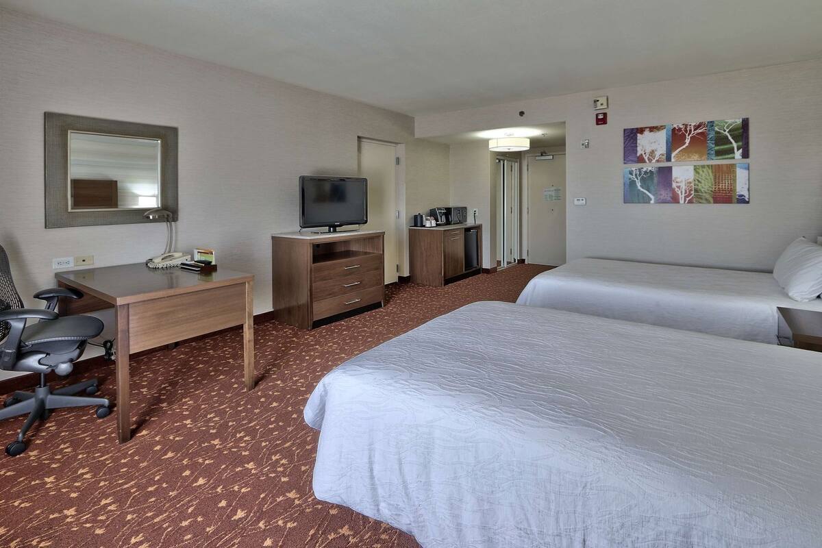 A hotel room with a desk, chair, two beds, a television, and a mirror.